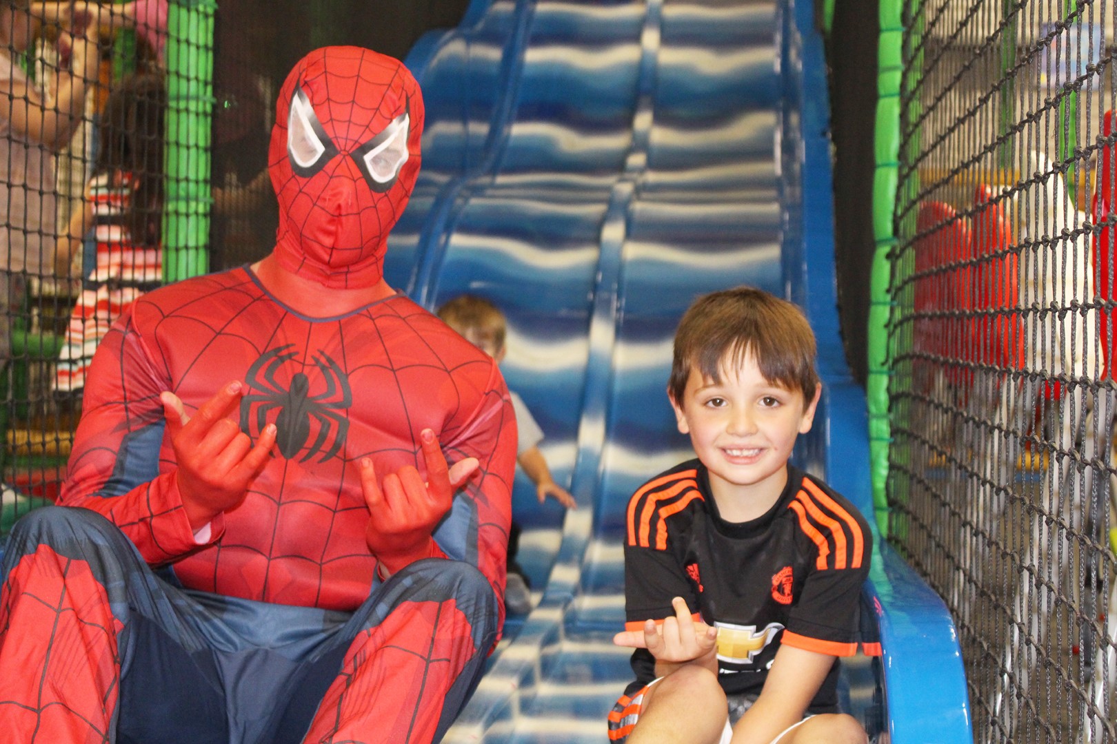 A smiling boy sitting with a Spider-Man character at the bottom of the big blue slide