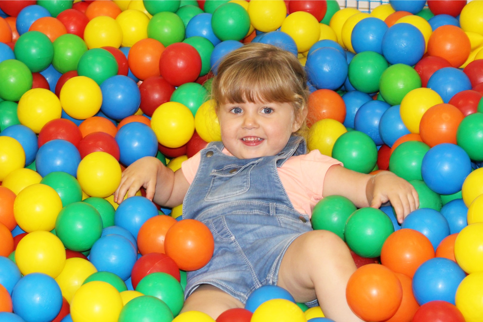 Little girl in denim dungarees smiling in a ball pit full of colourful balls