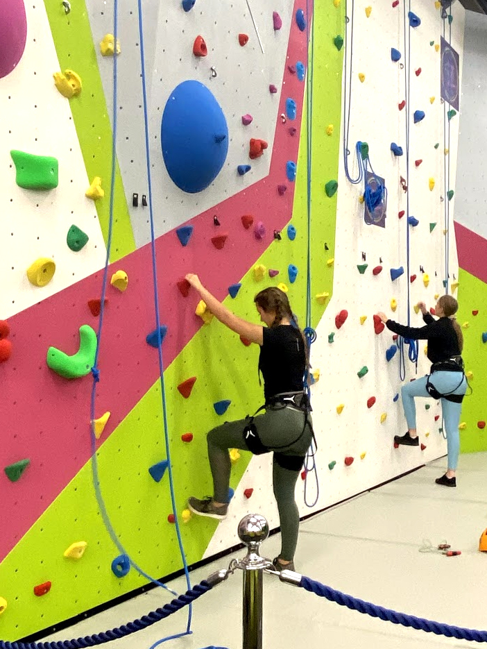 Two climbers in safety harnesses on the bright pink and lime climbing wall